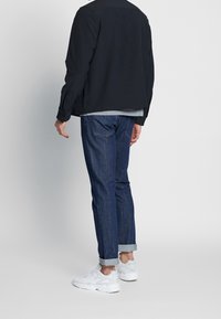 Sort jakke over en grå skjorte, parret med oprullede blå jeans og hvide sneakers. Glat stof og casual pasform med minimal dekoration.