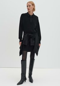 Vestito nero in maglia con collo a zip, abbinato a un cardigan con cintura e stivali in pelle fino al ginocchio. Texture liscia, design aderente.