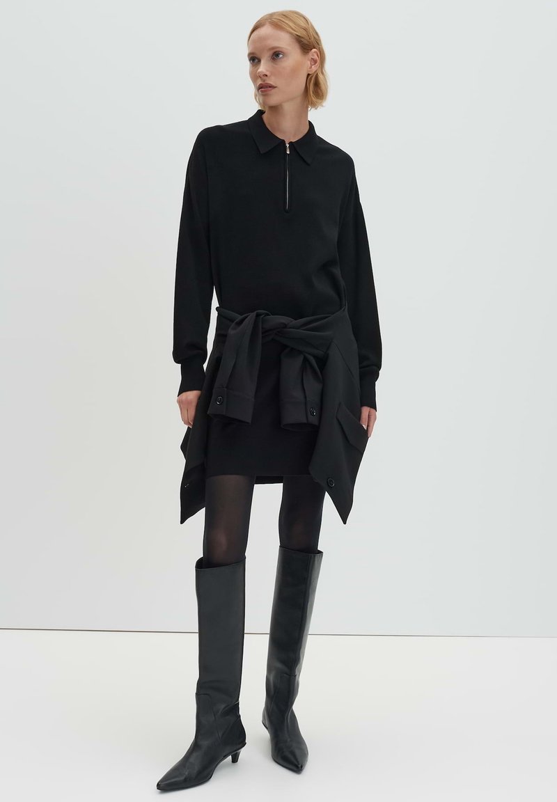 Vestito nero in maglia con collo a zip, abbinato a un cardigan con cintura e stivali in pelle fino al ginocchio. Texture liscia, design aderente.