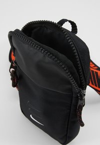 Černá crossbody taška ze tkaniny se zipovým uzávěrem, s nastavitelným popruhem a bílým logem na přední straně. Interiér podšitý jemným materiálem.