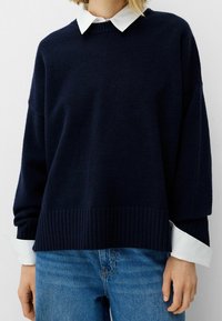 Pull bleu marine avec ourlet et poignets côtelés, portant une chemise blanche à col en dessous. Le tissu semble doux et légèrement texturé.