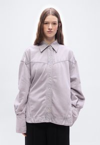 Νεαρή γυναίκα με μακριά μαλλιά, ντυμένη με ένα ανοιχτό μωβ φαρδύ σακάκι με κουμπιά και μαύρο παντελόνι, μπροστά σε απλό ανοιχτό φόντο.