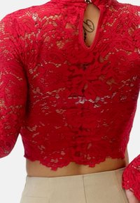 Top corto de encaje rojo con mangas largas y patrón floral, con cuello alto y un detalle de ojal en la parte trasera. Tejido texturizado.