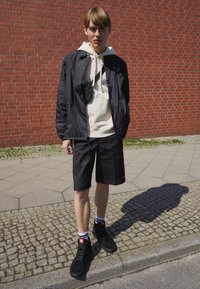 Veste légère noire sur un sweat à capuche crème, associée à un short noir et des chaussures montantes noires. Mur de briques texturé en arrière-plan.