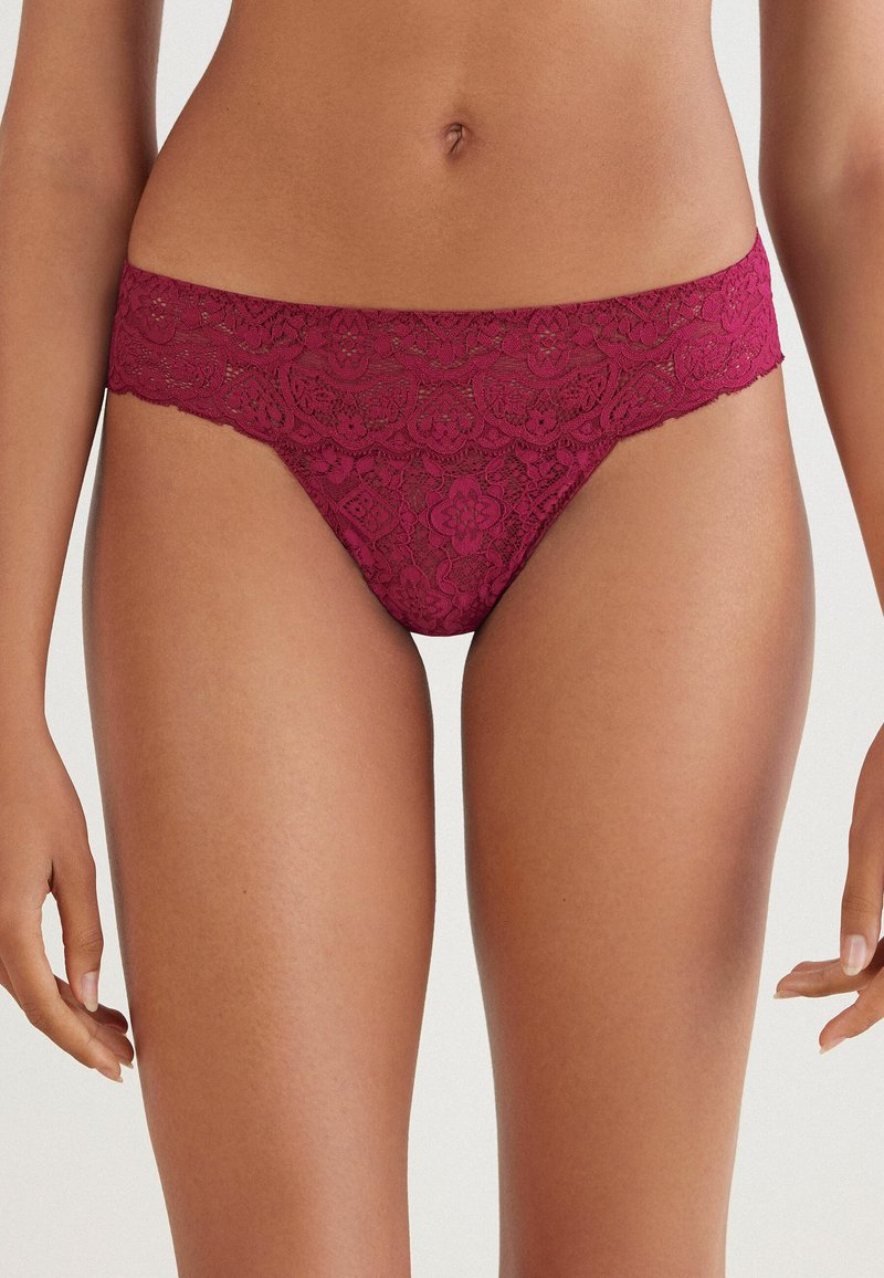 Mujer con ropa interior de encaje color burdeos estilo bikini, mostrando la zona del vientre y los muslos superiores sobre un fondo liso.