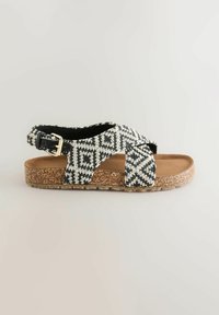 Sandale en tissu à motif noir et blanc avec semelle en liège, semelle intérieure couleur beige et bride réglable noire avec boucle, vue de côté sur un fond clair.