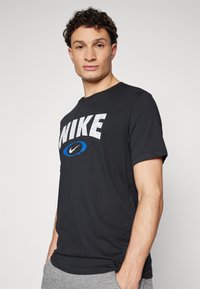 Crna pamučna majica s kratkim rukavima koja sadrži ispucani bijeli logo "NIKE" i plavi swoosh. Okrugli izrez, ležeran kroj.