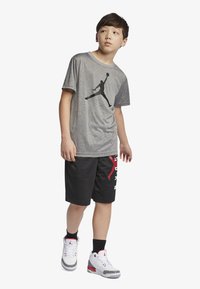 Grå t-skjorte med kort erme og svart silhuettlogo, paret med svarte nettshorts med rød og hvit tekst. Hvit sneakers med grå detaljer.