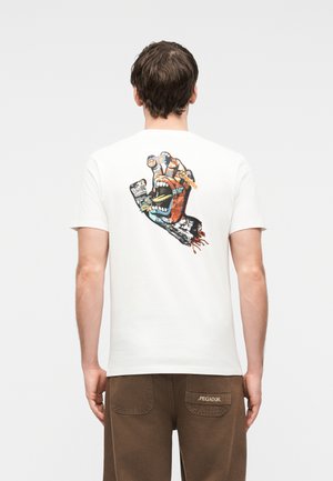 Bărbat purtând un tricou alb cu un grafic colorat pe spate, reprezentând o mână care țipă ținând un bilet, asortat cu pantaloni maro.