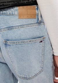 Los jeans de mezclilla azul claro presentan una etiqueta de cuero visible con la inscripción en relieve "TOMMY JEANS" y un pequeño logotipo tricolor en el bolsillo trasero.
