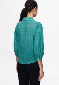 Mujer con cabello rizado recogido en una coleta, vistiendo una blusa a cuadros semi-transparente de color verde azulado con mangas abullonadas hasta el codo y pantalones negros, de espaldas.