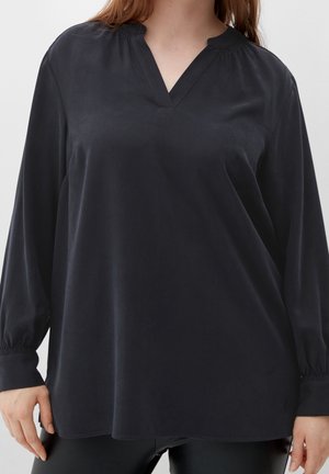 Žena v černé volné haleně s výstřihem do V, dlouhými rukávy s nadýchanými manžetami a elastickými lemy, v kombinaci s černými kalhotami, na jednobarevném pozadí.