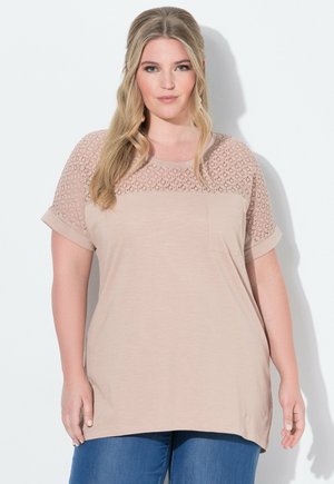 Model in een lichtbeige top met kantdetails op de mouwen, staand tegen een witte achtergrond, zelfverzekerd lachend met lang haar.