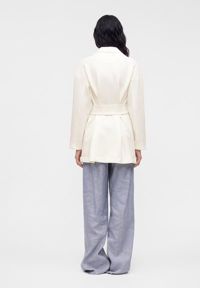 Femme aux longs cheveux foncés vue de dos, portant une veste blanche ceinturée et un pantalon ample gris clair sur un fond uni clair.