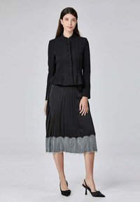 Veste noire ajustée avec fermeture à boutons, associée à une jupe plissée noire ornée d'une bordure en dentelle transparente grise. Portée avec des chaussures à talons noirs.