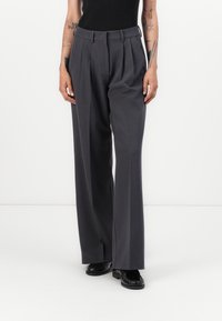 Wijde pantalon van donkergrijs stof, met geplooide voorkant, riemlussen en frontzakken. Gecombineerd met zwarte schoeisel.
