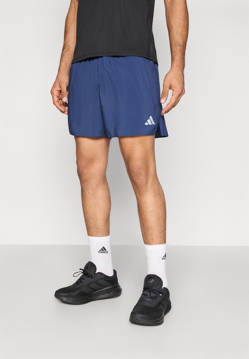 Marineblå treningsshorts laget av lett stoff, med sidespalte og hvit Adidas-logo. Kombinert med svarte løpesko og hvite sokker.