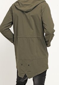 Giacca impermeabile verde oliva con cappuccio, caratterizzata da una vita arricciata, orlo asimmetrico e chiusure a pressione sul retro. Texture liscia.