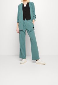 Teal kostuum met een blazer en wijde broekspijpen, laagjes over een zwart vest. De stof is glad, de broek heeft zijzakken en is gedragen met witte sneakers.