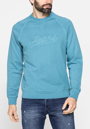 Uomo con la barba che indossa una felpa verde acqua ricamata con "Jeans" e jeans di denim blu scuro consumati su uno sfondo semplice.