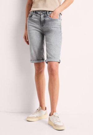 Person iført lysegrå opkørte denimshorts, beige top og hvide sneakers, står foran en ensfarvet lys baggrund.