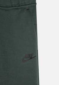 Olivno zeleni Nike trenirke, ki imajo črn logotip in šivske detajle. Narejene so iz mehkega materiala z teksturirano površino.