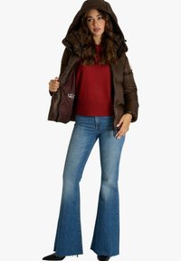 Giacca a puff marrone con cappuccio, chiusura a zip e consistenza lucida, indossata sopra un maglione rosso; abbinata a jeans a zampa di elefante e scarpe nere.