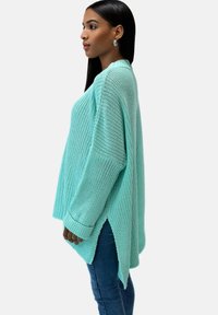 Türkiser lockerer Strickpullover mit seitlichen Schlitzen, oversized Schnitt, gerippten Bündchen und strukturiertem Stoff, kombiniert mit Jeans.