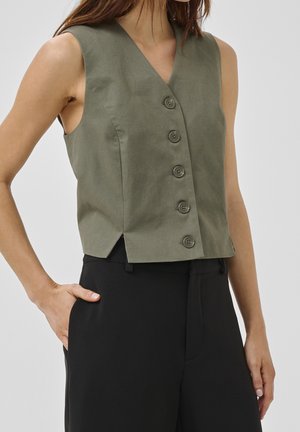 Femme portant une veste sans manches olive boutonnée et un pantalon noir taille haute, une main dans la poche sur fond uni.