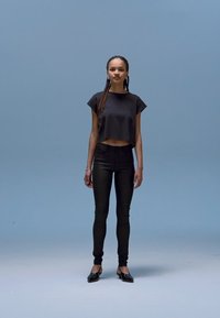 Top curto preto, calças pretas de cintura alta, sapatilhas pretas. Em pé sobre um fundo azul claro, cabelo liso, acessórios mínimos.