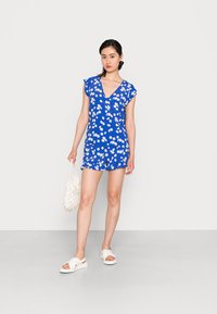 Macacão floral azul com mangas curtas e decote em V. Apresenta padrões de margaridas brancas e uma barra solta. Combinado com sandálias brancas e uma mala texturizada.