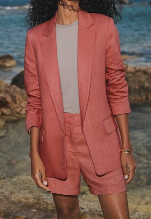 Textured, coral pink suit with a tailored blazer and matching shorts. The blazer features a single button and side pockets, paired with a gray top.