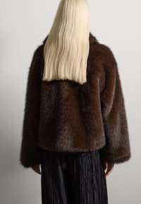 Chaqueta de piel sintética marrón con una textura suave y esponjosa y mangas anchas, combinada con pantalones negros plisados. Vista posterior mostrada contra una pared.