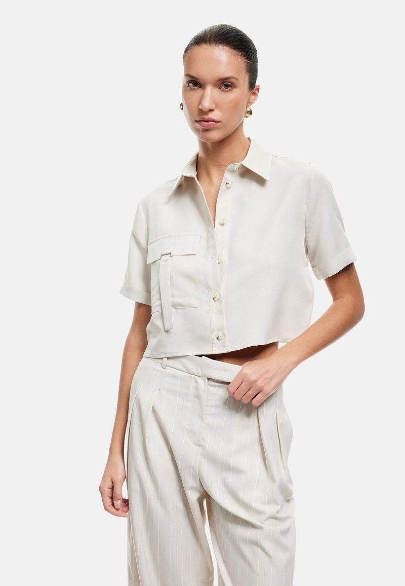 Blusa di crema tagliata con maniche corte, grande tasca sul petto e chiusura a bottoni, abbinata a pantaloni a righe sottili di colore chiaro.