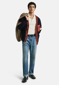 Marineblauer Cardigan mit orangen Streifen über einem weißen Poloshirt, kombiniert mit hellblauen Jeans und schwarzen Schuhen, hält ein kariertes Sakko.