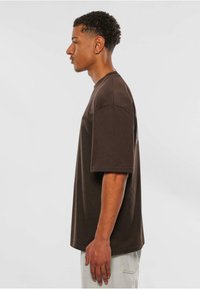 Brun bomull oversized t-skjorte med korte ermer, rund hals, synlige sømmer og logo detalj. Modellen står i profil mot en enkel bakgrunn.