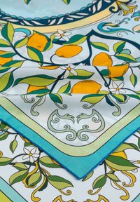 Seidenstoff mit Zitronen- und grünen Blätter-Muster, blauen und gelben dekorativen Rändern und weißen Blumen, leicht gefaltet.