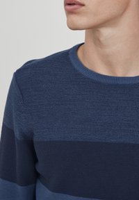 Mėlynas megztinis su įvairiais tekstūriniais dryžiais. Yra apvalia iškirpte ir siauro profilio dizainu.