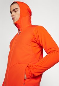 Bright orange zip-up jacket with a high collar and hood, featuring textured sleeves and side pockets, made from lightweight material.