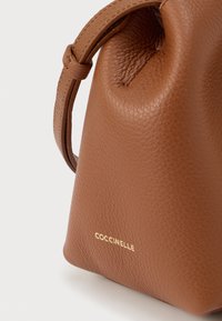 Sac à main en cuir marron texturé avec bandoulière réglable et logo doré "COCCINELLE" embossé dans le coin inférieur avant.