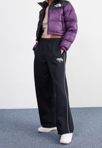 Person wearing cropped purple North Face jacket, black Nike sweatpants with white piping, and white sneakers standing on blue carpet.