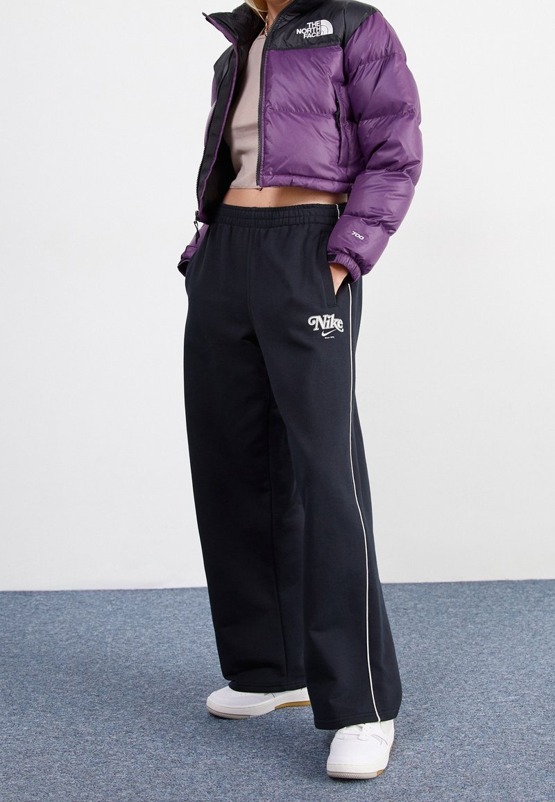 Person wearing cropped purple North Face jacket, black Nike sweatpants with white piping, and white sneakers standing on blue carpet.