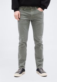 Άτομο που φοράει γκρι τζιν στενής γραμμής, μαύρα αθλητικά παπούτσια με λευκά κορδόνια, μαύρο φούτερ, στέκεται με τα χέρια στις τσέπες σε απλό φόντο.