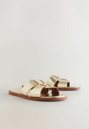 Sandali metallici dorati con ampiezza incrociata, design con punta aperta e suola piatta marrone. Texture liscia con un'estetica minimalista.