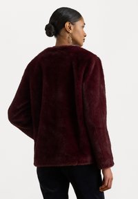 Burgundi színű műszőrme dzseki laza fazonnal, hosszú ujjak és puha textúra, hátulról látható, sima fehér háttér előtt.