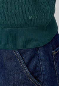 Pull en tricot texturé vert avec ourlet côtelé, arborant un logo BZB discret. Associé à un jean en denim bleu foncé, avec des coutures jaunes.