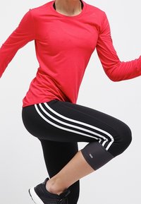 Osoba w czerwonej koszulce z długim rękawem i czarnych legginsach z białymi paskami, podnosząca jedno kolano, ubrana w czarne buty sportowe na jednolitym tle.