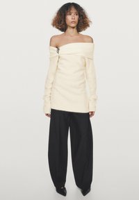 Kvinde med krøllet hår iført en off-shoulder creme sweater med en broche, brede sorte bukser og sorte spidse sko, stående mod en hvid baggrund.