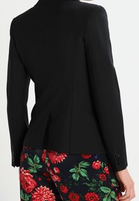 Zwarte op maat gemaakte blazer met een getailleerd ontwerp, een gladde textuur en een enkele split aan de achterkant, gecombineerd met kleurrijke bloemenprintbroeken.