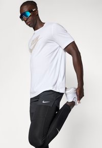 Fehér Nike póló "RUN" grafikával, fekete Nike sportnadrággal, fekete leggingssel és fehér sportcipővel. A téma nyújtja a lábát, miközben kék napszemüveget visel.